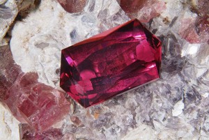 Faceted Rubellite (red tourmaline) from Morro Redondo, Minas Gerais, Brazil; sitting on a piece of pegmatite containing rough rubellite crystals, quartz and lepidolite mica.